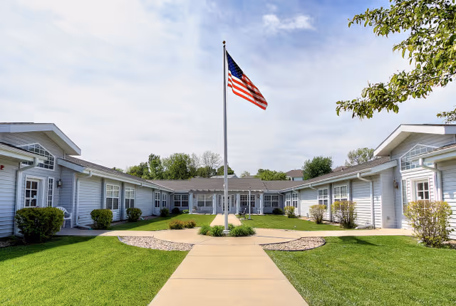 Courtyard entrance of a single-story senior living building with a central American flagpole, paved walkway, and manicured lawns.