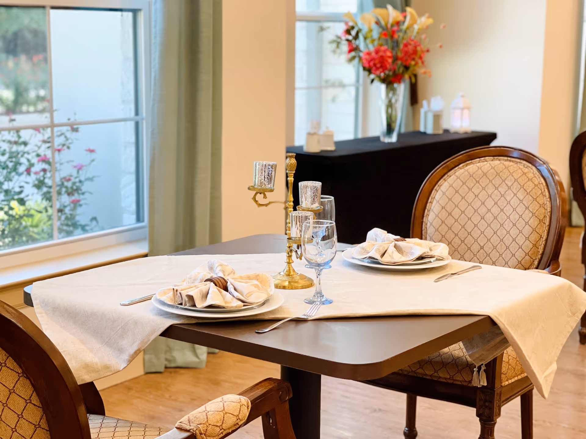 A dining table set for two with beige napkins on white plates, silverware, and clear water glasses. The table has a cream-colored tablecloth and a gold candelabra centerpiece. The background shows a window with green curtains and a side table with a vase of colorful flowers.