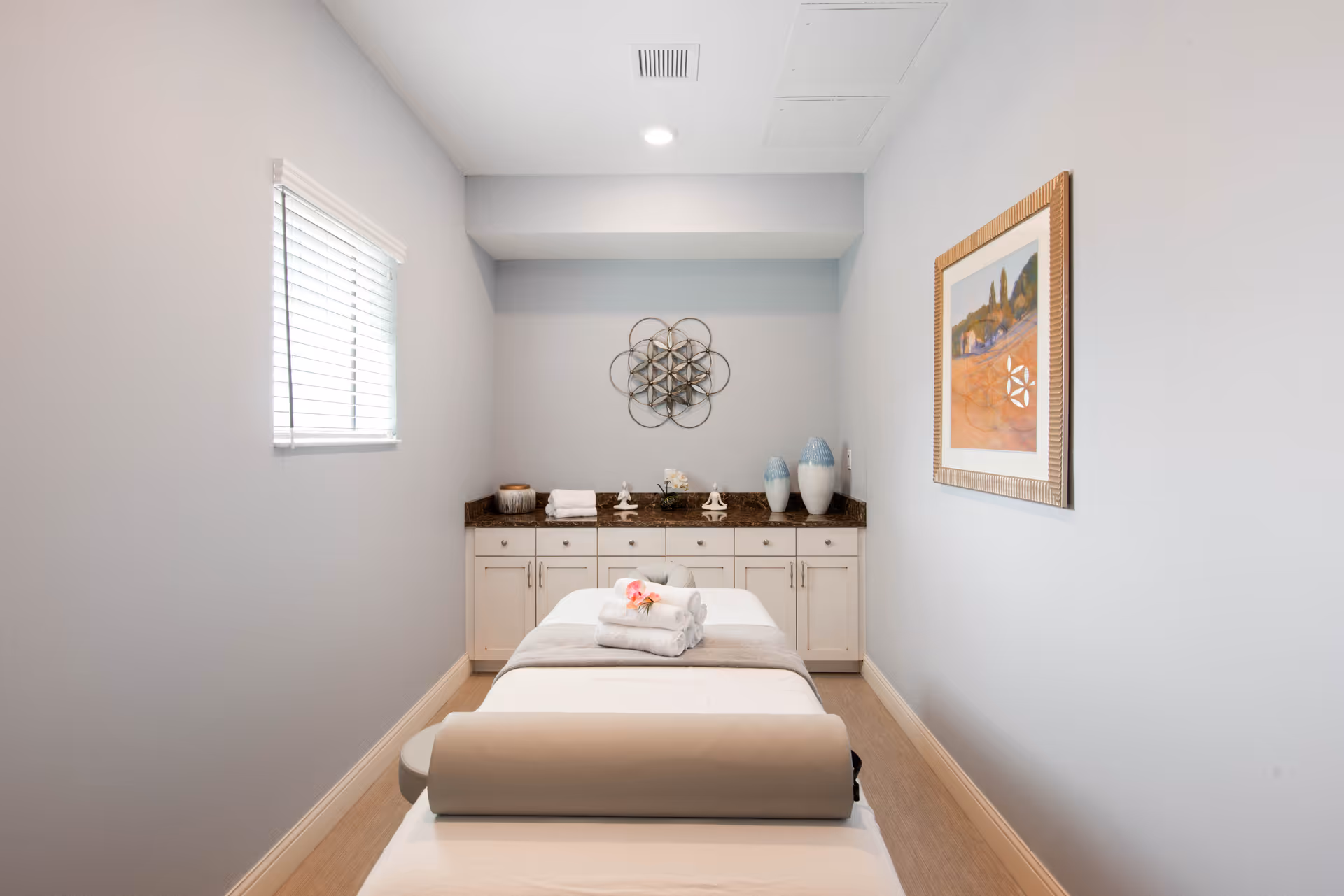 A small, serene massage therapy room with a massage table in the center. The table is covered with white sheets and has neatly folded towels and a pink flower on top. The room has light gray walls, a small window with blinds on the left, a countertop with cabinets underneath at the back, decorative vases, and a framed painting on the right wall.
