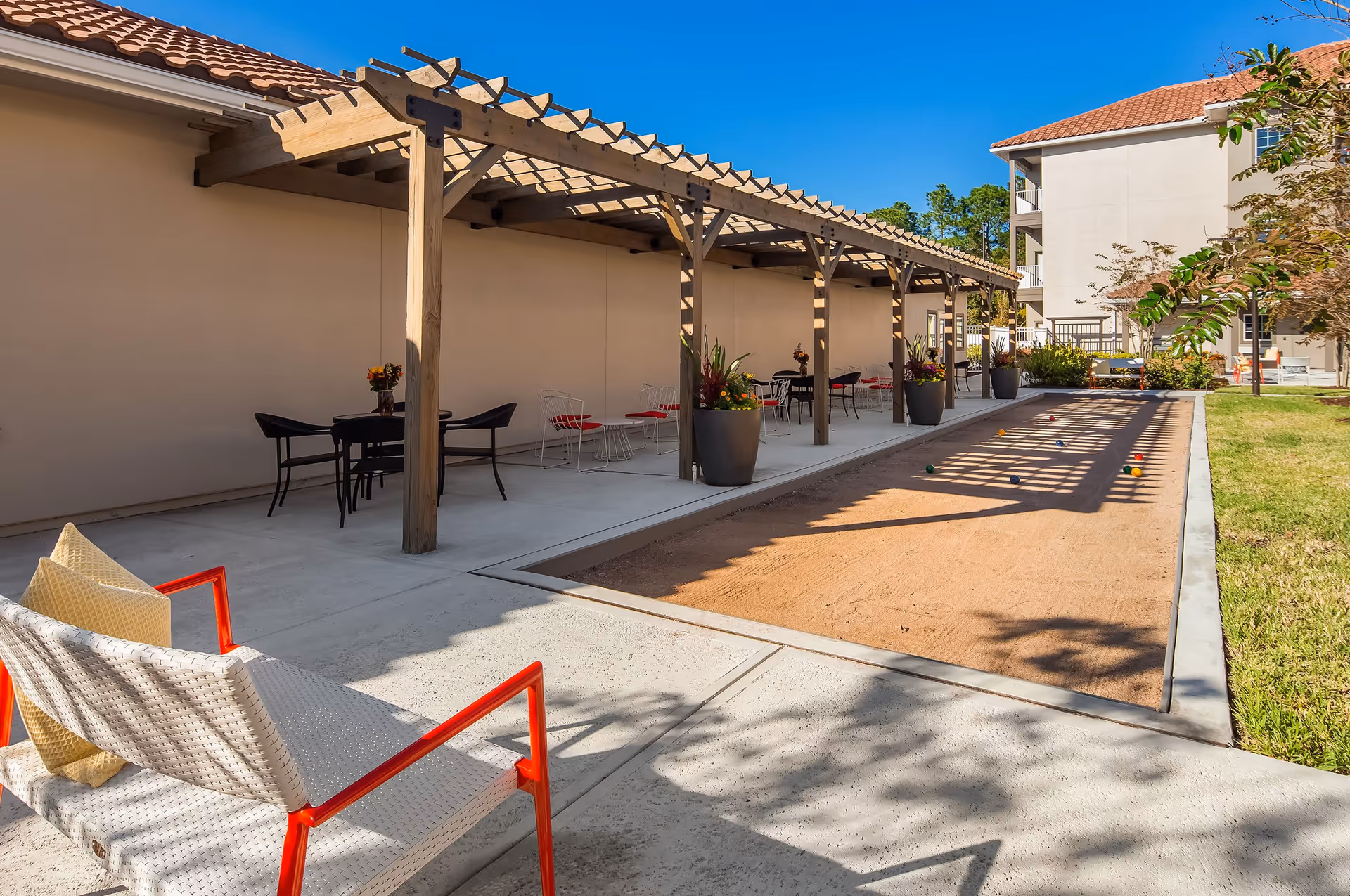 Outdoor bocce ball court with a wooden pergola providing shade over several tables and chairs. There are potted plants along the pergola and a white bench with red armrests and yellow cushions in the foreground. The scene is set in a sunny courtyard with a building in the background.