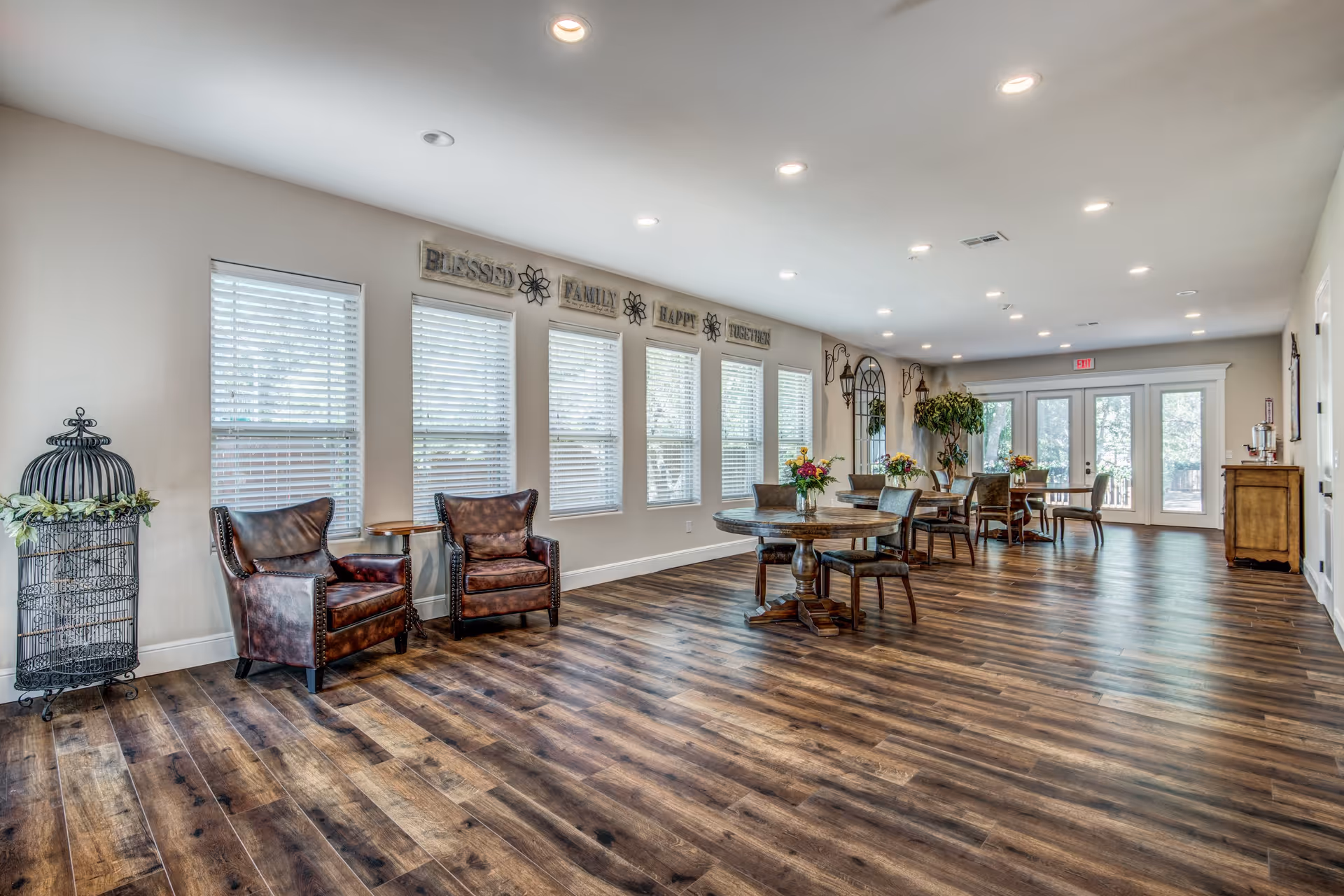 Spacious, well-lit communal living and dining area with wooden floors, leather chairs, round tables, and large windows.