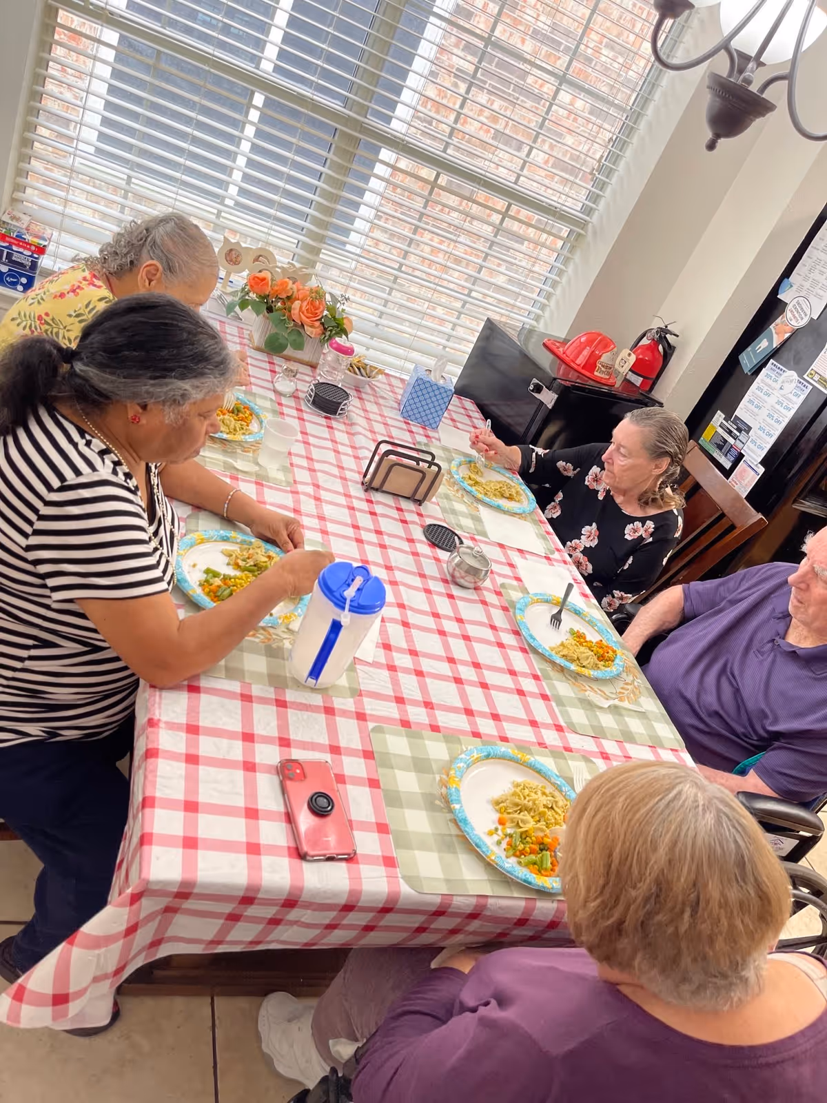 Five elderly individuals sitting around a dining table with a red and white checkered tablecloth, eating a meal consisting of pasta and vegetables. The room is well-lit with natural light coming through large windows with blinds. There are flowers and various items on the table, and a fire extinguisher and red fire hat are visible in the background.