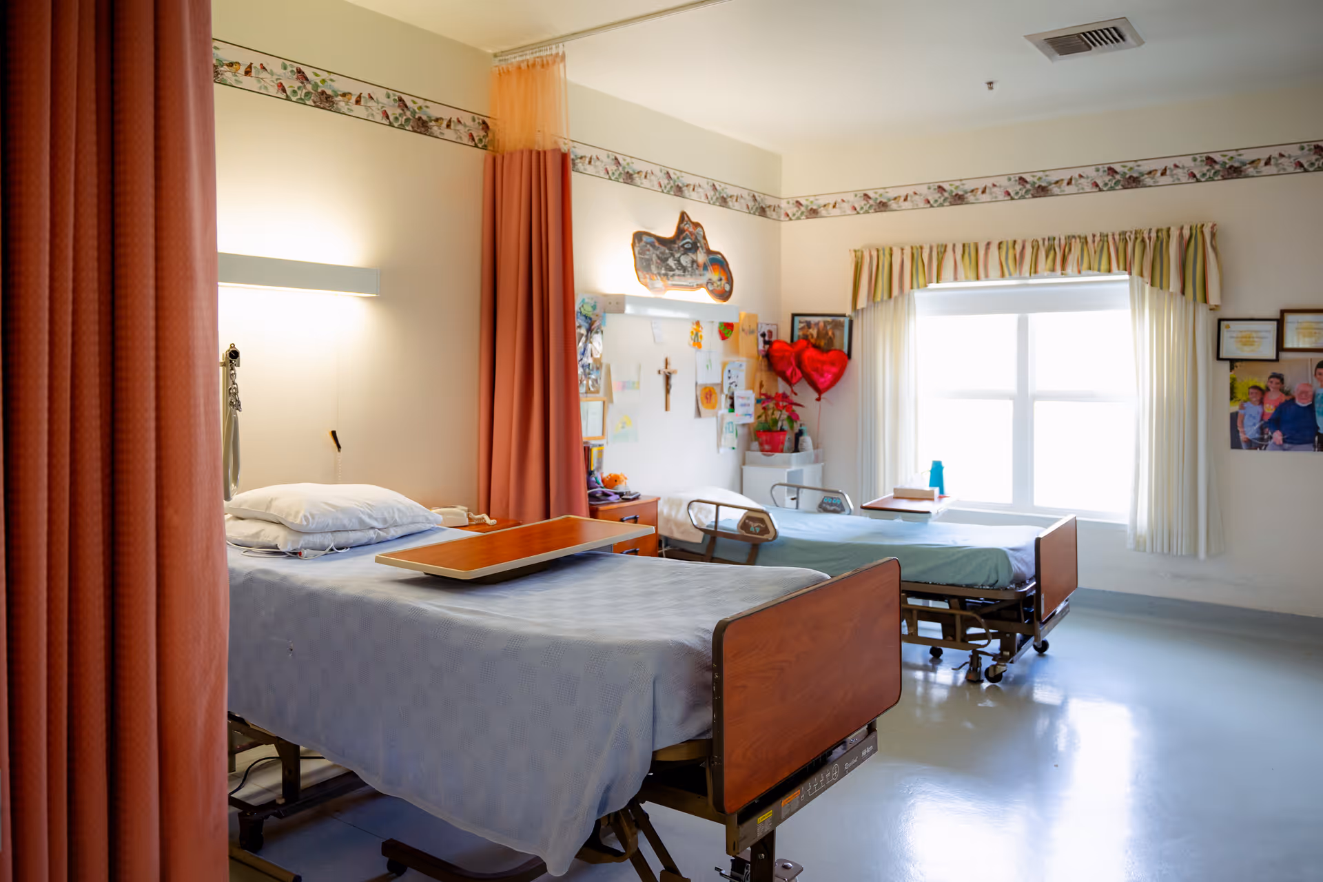 A bright room with two hospital-style beds separated by a curtain. Each bed has a wooden headboard and footboard, with a tray table on one bed. The walls are decorated with a floral border near the ceiling, various pictures, and two red heart-shaped balloons near the window with striped curtains. The floor is smooth and light-colored.