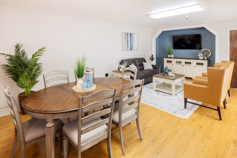 A bright and cozy living room and dining area with wooden flooring. The dining area features a wooden table with six cushioned chairs and decorative plants. The living room has a dark gray sofa, two tan armchairs, a white coffee table, a white media console, and a wall-mounted flat-screen TV. The walls are light-colored with a blue accent wall behind the TV, and there is a framed abstract painting on the wall.