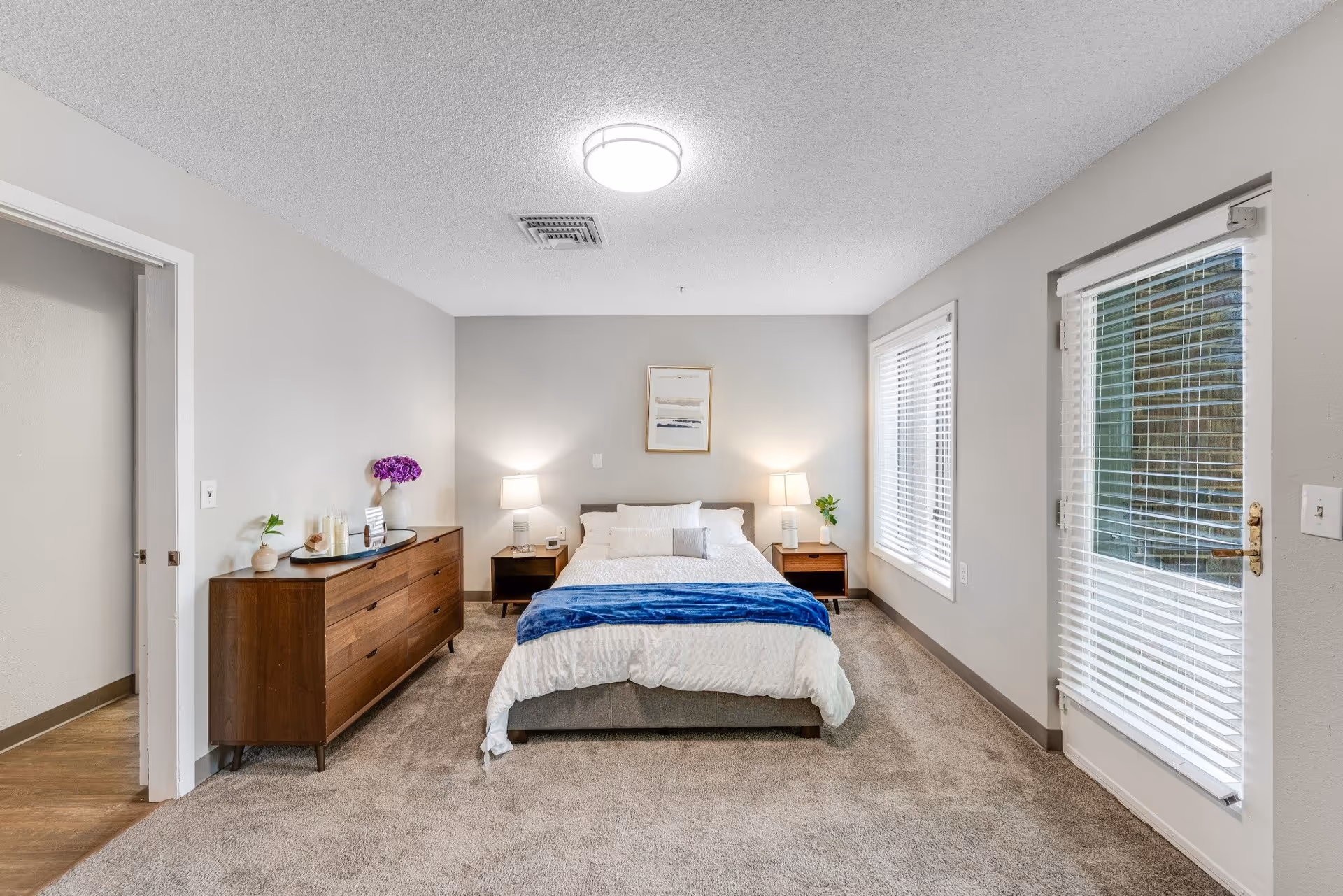 Bright, neatly staged bedroom with a bed centered between nightstands, a wooden dresser to the left, and a glass door with blinds to the right.