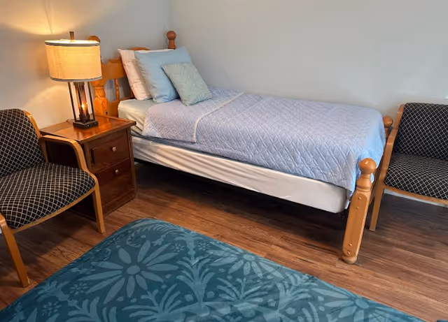 A furnished senior living bedroom with single bed, nightstand, lamp, and two chairs.