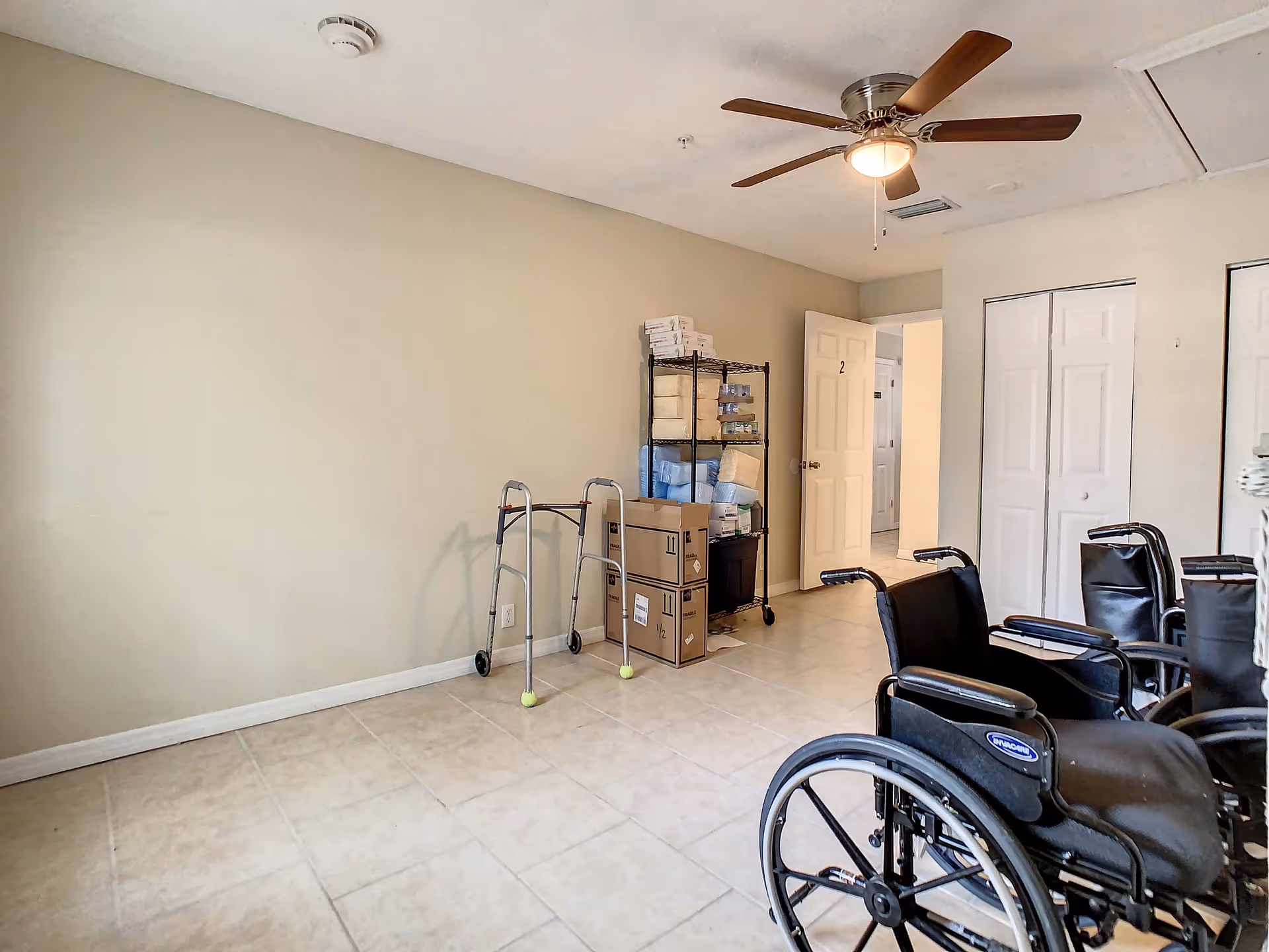 A room with beige walls and tiled floor containing several black wheelchairs on the right side, a metal shelving unit with medical supplies and boxes, and a walker with tennis balls on the front legs. There is a ceiling fan with wooden blades and an open door labeled with the number 2 leading to a hallway.