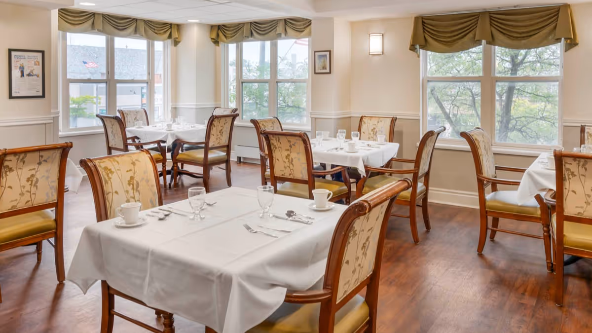 Bright dining room with white-clothed tables and upholstered wooden chairs arranged near large windows.