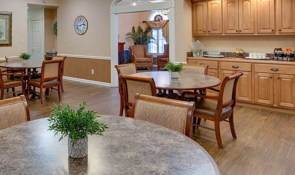 A dining room with round tables and chairs, small potted centerpieces, and a kitchenette with wooden cabinets in a senior living facility.