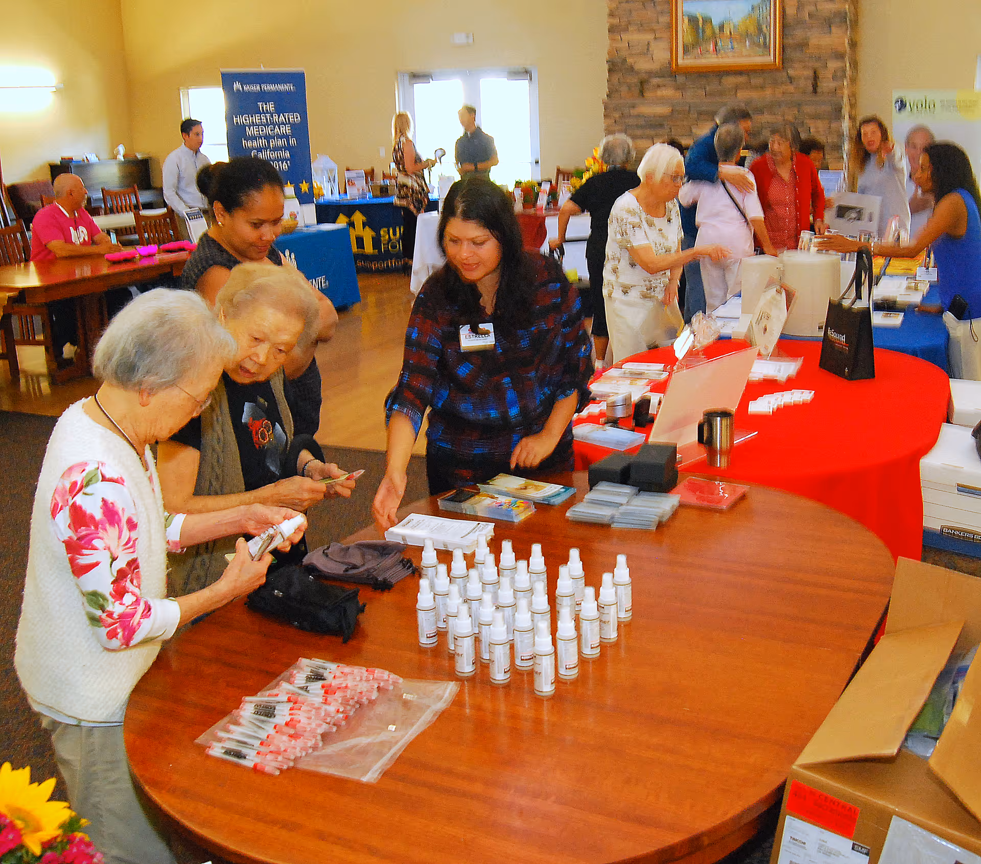 A group of elderly people and staff interacting at a community event inside a senior living facility. Several tables are set up with informational brochures, small bottles, and other items. People are engaged in conversation and examining the materials on the tables. The room has warm lighting and a welcoming atmosphere.