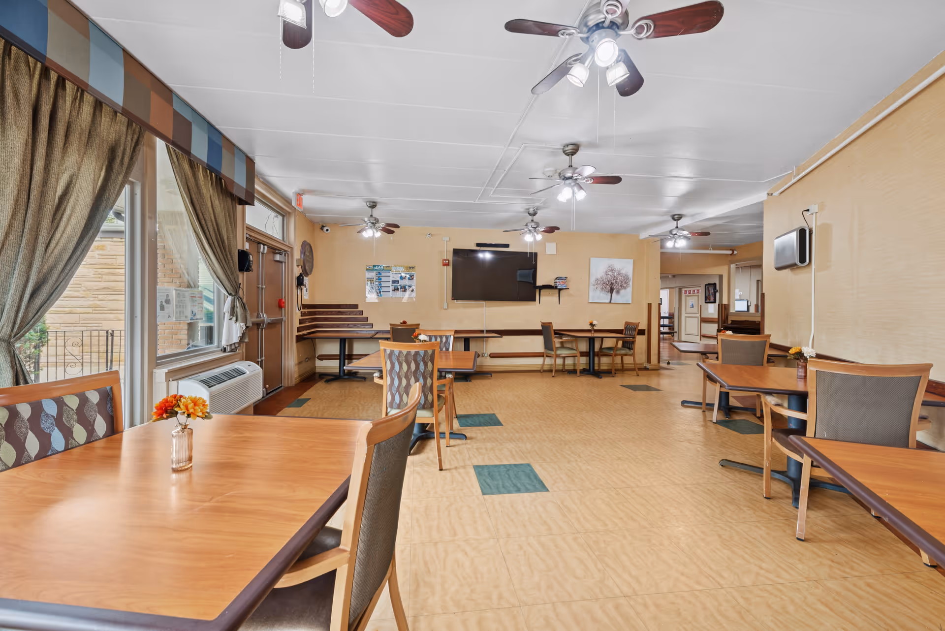 Spacious communal dining room with wooden tables and chairs, ceiling fans, a wall-mounted TV, and large curtained windows.