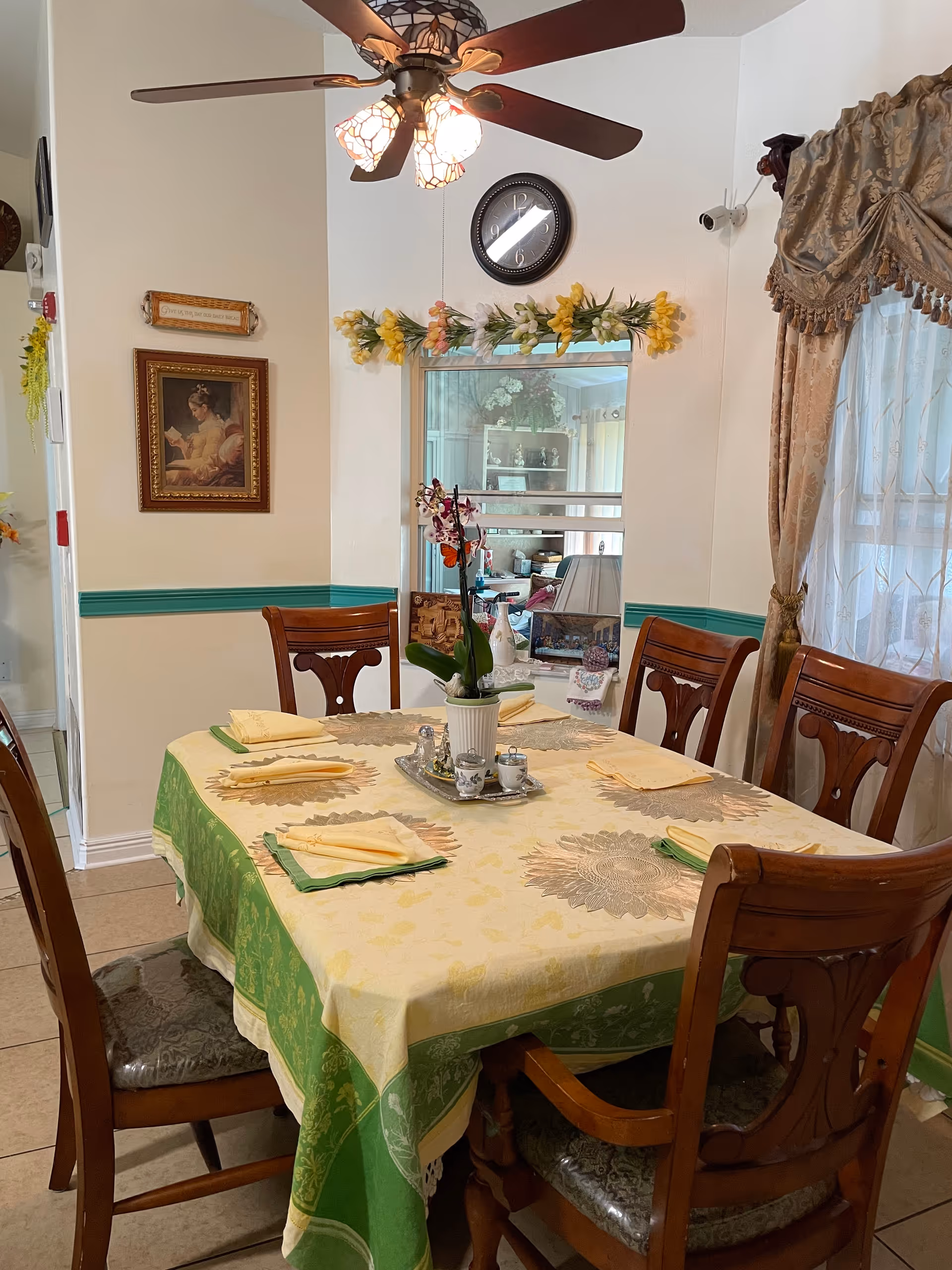 A dining room with a rectangular wooden table covered with a yellow and green tablecloth, set with six folded yellow napkins. Six wooden chairs with cushioned seats surround the table. A ceiling fan with lights hangs above. On the wall, there is a clock, a floral garland, a framed painting of a woman reading, and a window with curtains. The room has a cozy and traditional decor style.