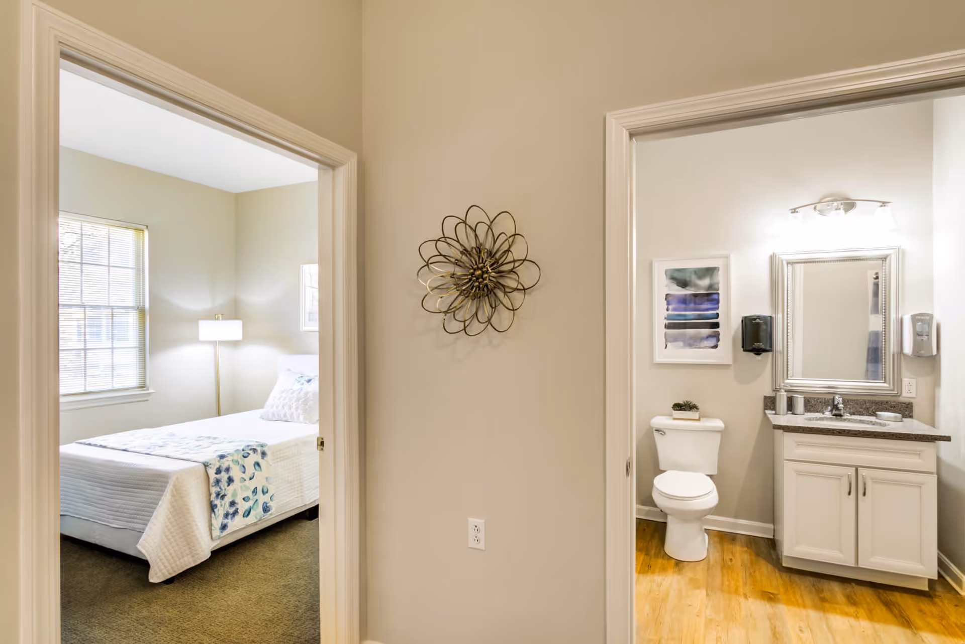 View of a hallway with two open doorways, one leading to a bedroom with a bed, a floor lamp, and a window, and the other leading to a bathroom with a toilet, a vanity with a sink and mirror, wall-mounted soap dispensers, and framed artwork.