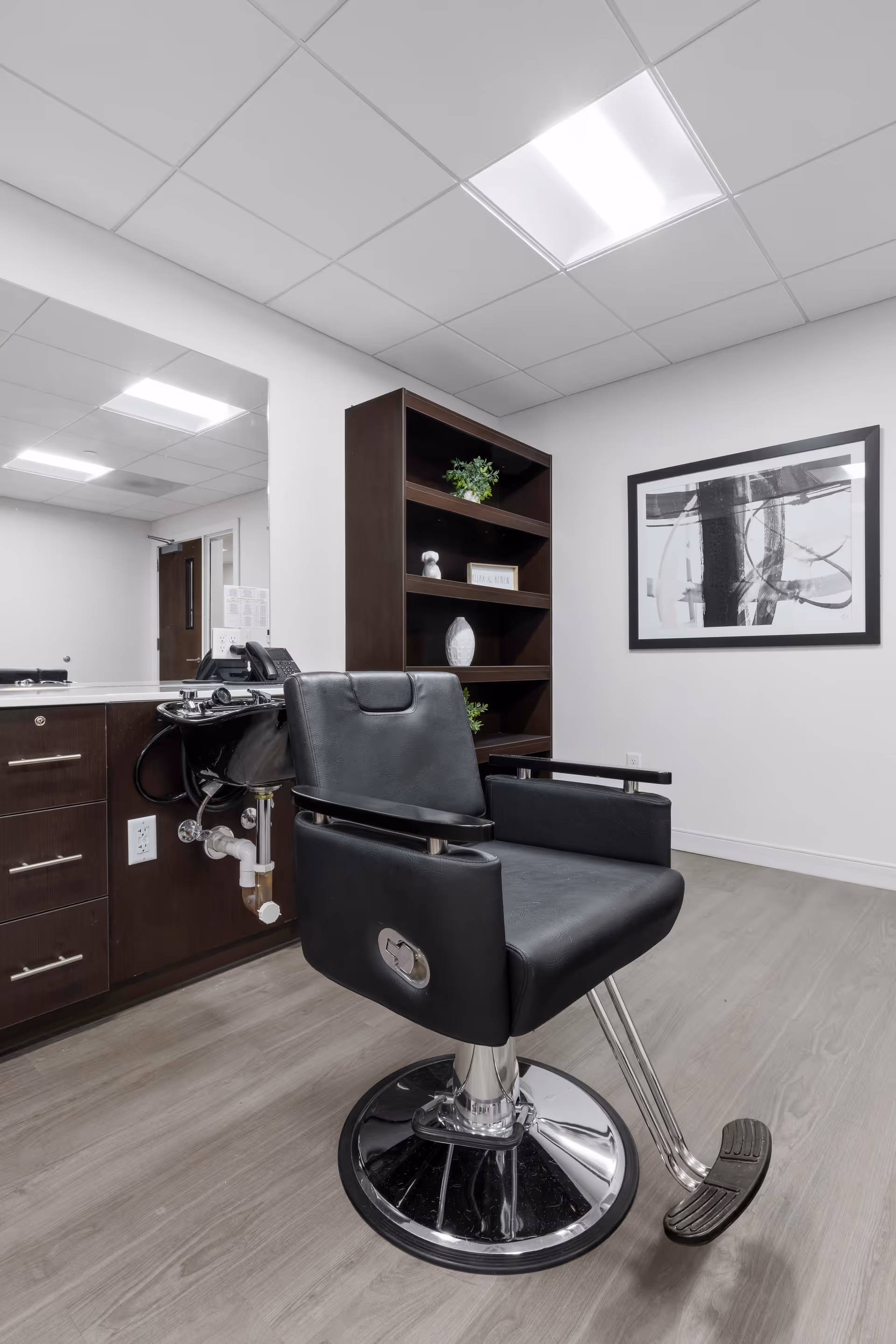 Interior view of a salon or barber area in a senior living facility. The image shows a black salon chair with a footrest in front of a dark wood cabinet with drawers and a sink. Behind the chair is a dark wood shelving unit with decorative items and a framed abstract black and white artwork on the wall. The floor is light wood, and the ceiling has recessed lighting panels.