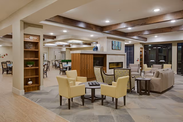 A spacious and well-lit senior living common area featuring a central fireplace surrounded by comfortable seating including beige and patterned armchairs. There are tables with decorative items, wooden beams on the ceiling, and a dining area with tables and chairs visible in the background.