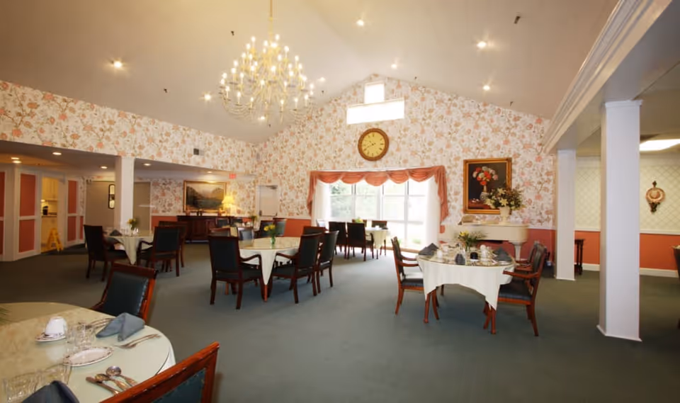 A spacious dining room with several round tables covered with white tablecloths and set with plates, glasses, and napkins. The room features floral wallpaper, a large chandelier hanging from a vaulted ceiling, a clock above a window with draped curtains, and framed artwork on the walls. There are multiple chairs around each table and a piano with a flower arrangement on top.