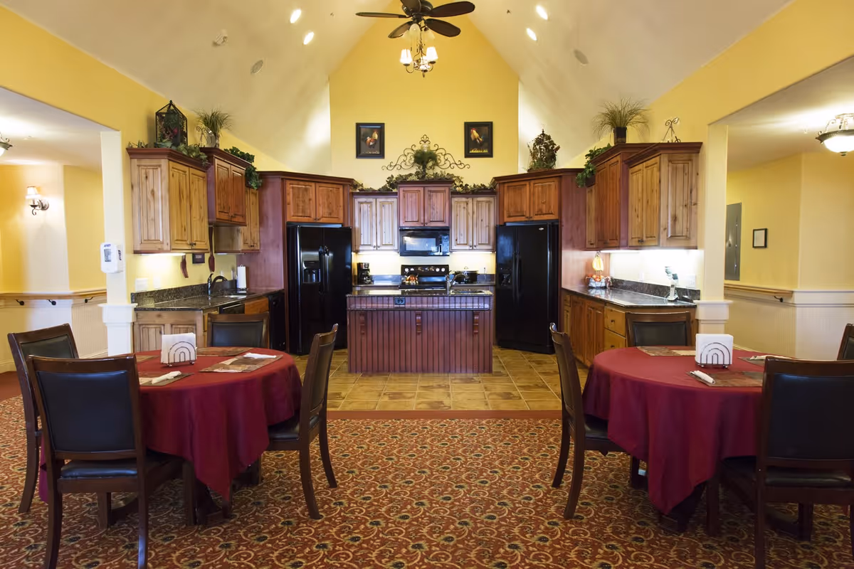Interior view of a kitchen and dining area in an assisted living facility. The kitchen features wooden cabinets, two black refrigerators, a black microwave, and a central island. The dining area has round tables covered with red tablecloths and surrounded by dark wooden chairs with black cushions. The walls are painted yellow, and the ceiling is vaulted with a ceiling fan and chandelier. Decorative plants and framed pictures adorn the top of the cabinets.