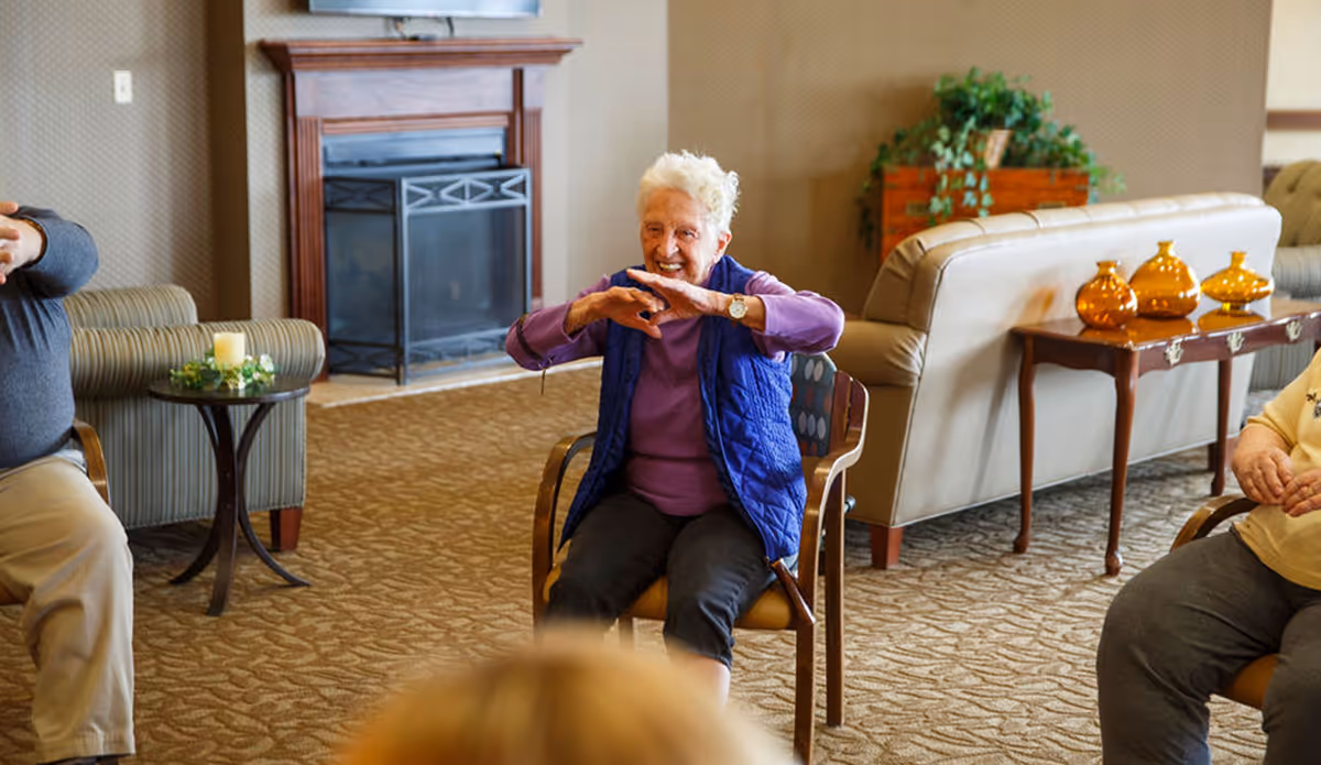 An elderly woman sitting on a chair in a senior living facility common area, smiling and stretching her arms. The room has a fireplace, comfortable seating, a small table with a candle, and decorative plants and vases.