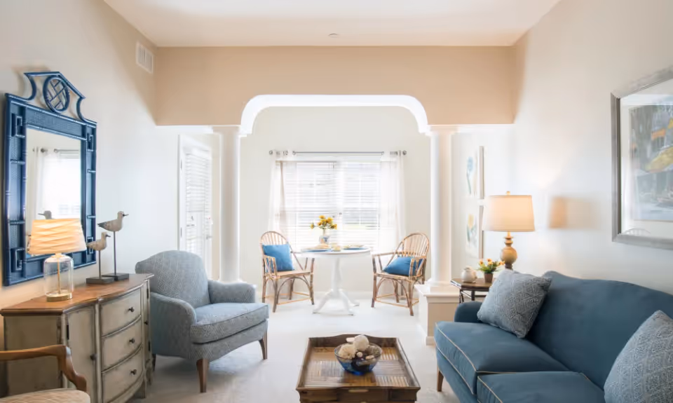 Bright, softly decorated living room with a blue sofa and armchair, wooden coffee table, sideboard, and a small sunlit breakfast nook by a window.