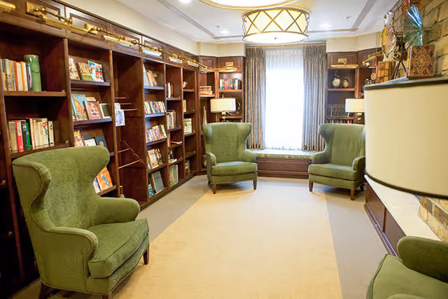 A cozy library or reading room with wooden bookshelves filled with books along the walls, three green upholstered wingback chairs arranged around a beige carpet, a window with curtains letting in natural light, and a ceiling light fixture.