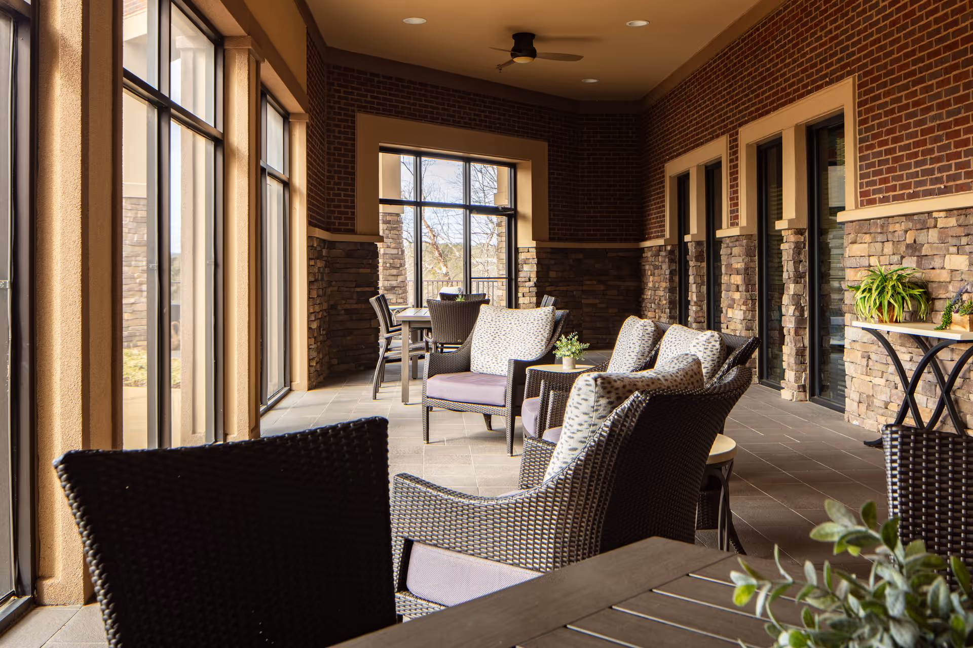 Covered outdoor seating area with wicker chairs, cushioned armchairs, tables and potted plants along a brick and stone wall with large windows.