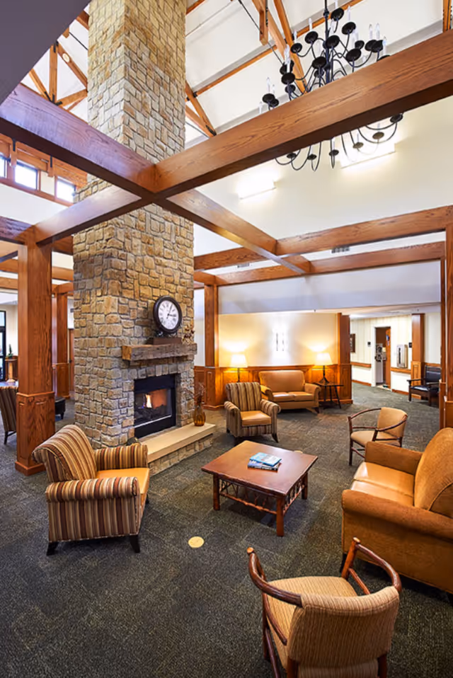A cozy living room area in a senior living facility featuring a large stone fireplace with a clock on the mantel, surrounded by wooden beams and a high ceiling. The room is furnished with various armchairs and sofas arranged around a wooden coffee table with books on it. Warm lighting from lamps and wall sconces creates a welcoming atmosphere.