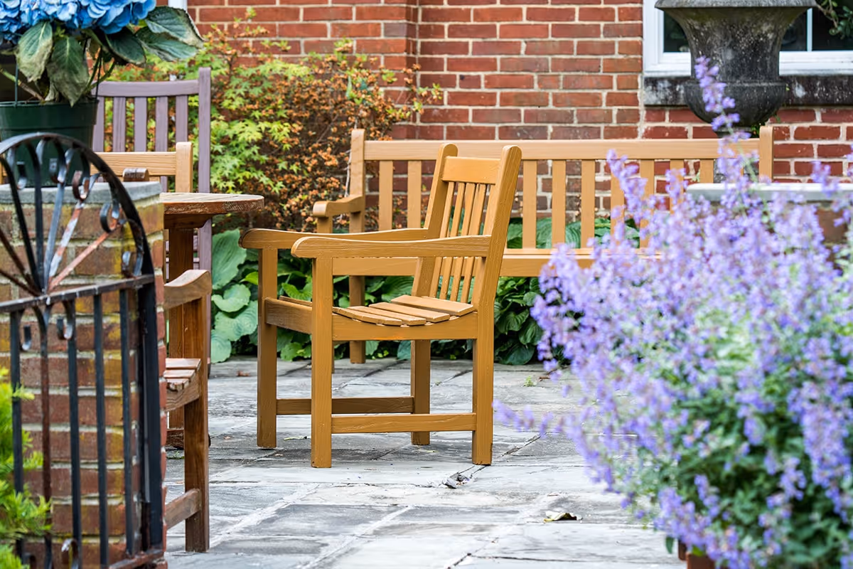 Wooden benches and chairs on a stone patio beside a brick wall with flowering plants.