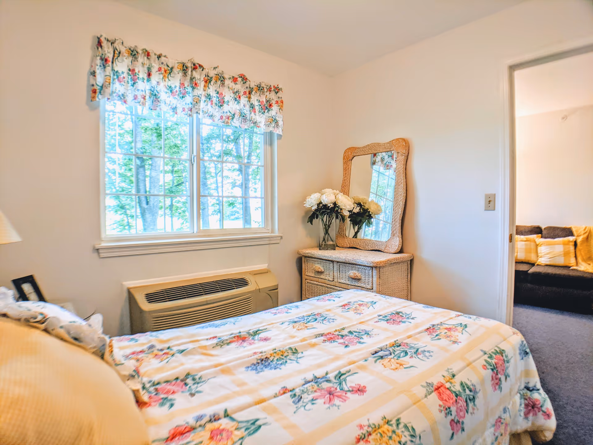 A bright bedroom with a floral bedspread, a window, wicker dresser with mirror and a vase of flowers.