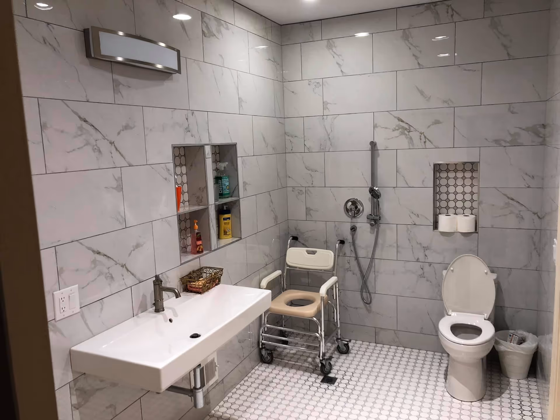 A senior-friendly bathroom with marble-patterned wall tiles and hexagonal floor tiles. The room features a wall-mounted sink with a modern faucet, a shower area with a handheld showerhead, a white toilet with the lid open, and a shower chair with wheels. There are built-in shelves holding toiletries and toilet paper rolls, and a small trash bin next to the toilet.