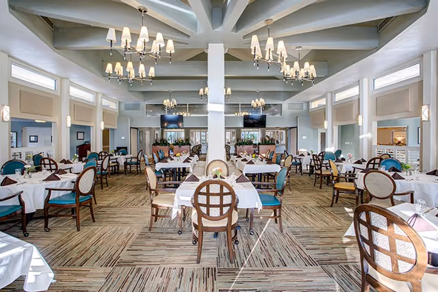 Spacious, well-lit dining room with multiple tables set with white tablecloths and chairs beneath chandeliers.