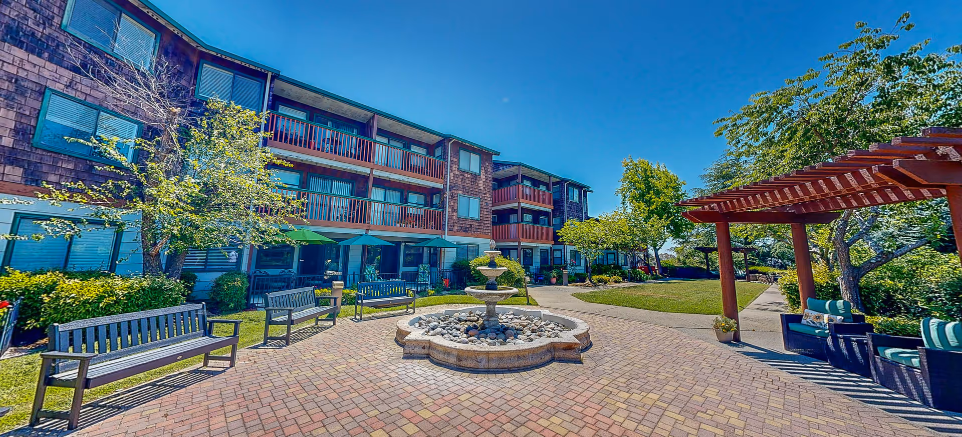 Outdoor courtyard area of a senior living facility with a central stone fountain surrounded by benches and patio umbrellas. The three-story building with balconies and windows is visible in the background, along with trees and a pergola with seating on the right side under a clear blue sky.