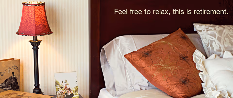 A cozy bedroom corner featuring a bed with white and rust-colored pillows, a dark wooden headboard, and a bedside table with a decorative lamp and framed photos. Text on the image reads, 'Feel free to relax, this is retirement.'
