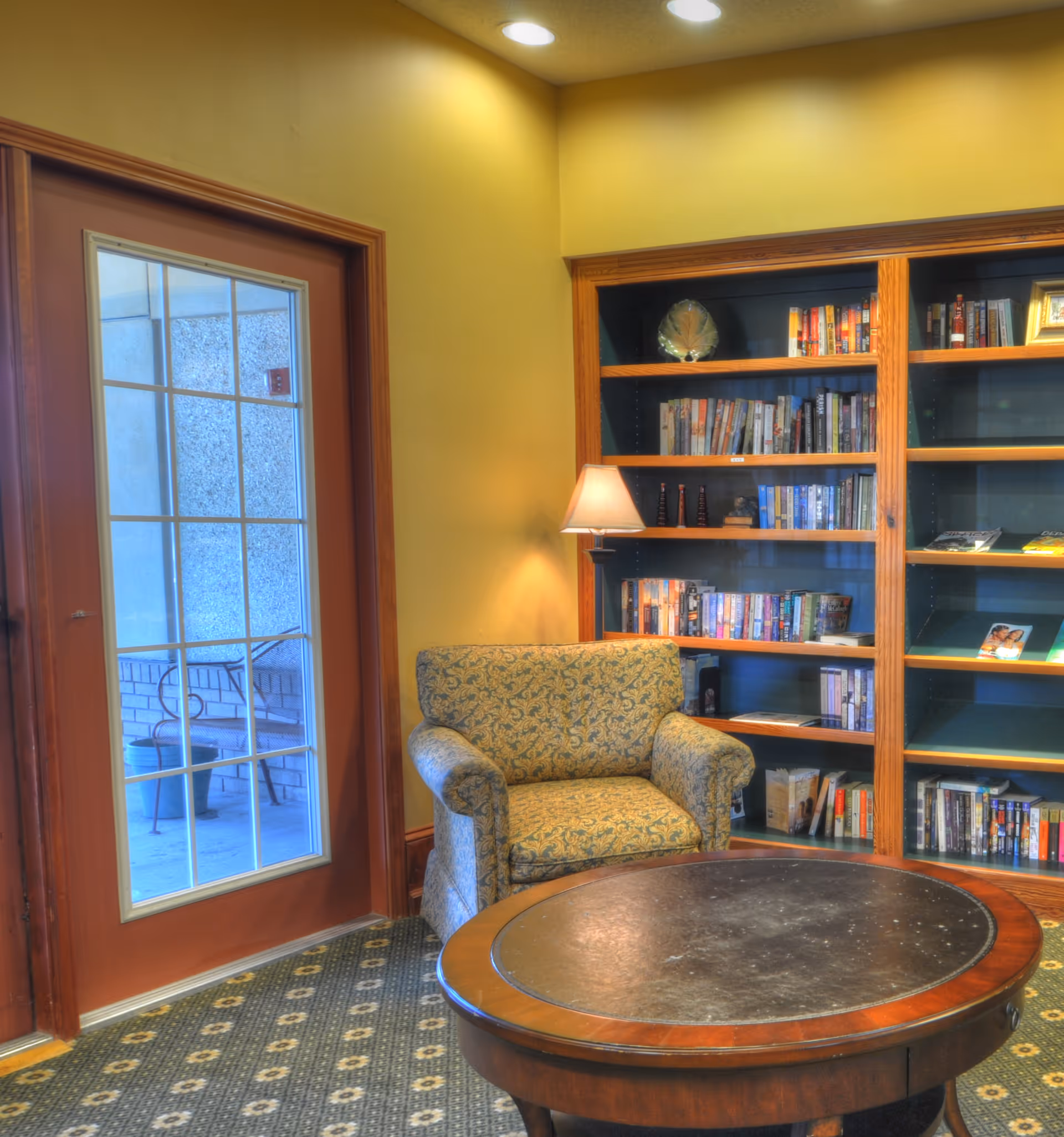 A cozy reading nook featuring a patterned armchair, a round wooden coffee table, and a built-in bookshelf filled with books and decorative items. A floor lamp provides warm lighting, and a glass door leads to an outdoor area with a bench.