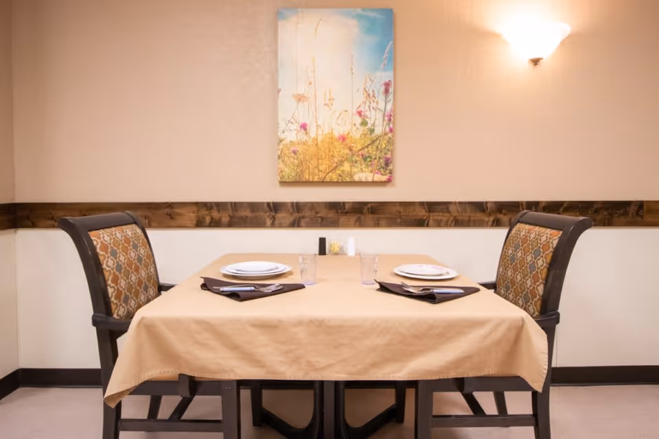 A small dining table set for two with a beige tablecloth, two patterned cushioned chairs, two plates, two glasses, and napkins with cutlery. The wall behind the table has a wooden trim, a light fixture, and a colorful painting of wildflowers and a blue sky.