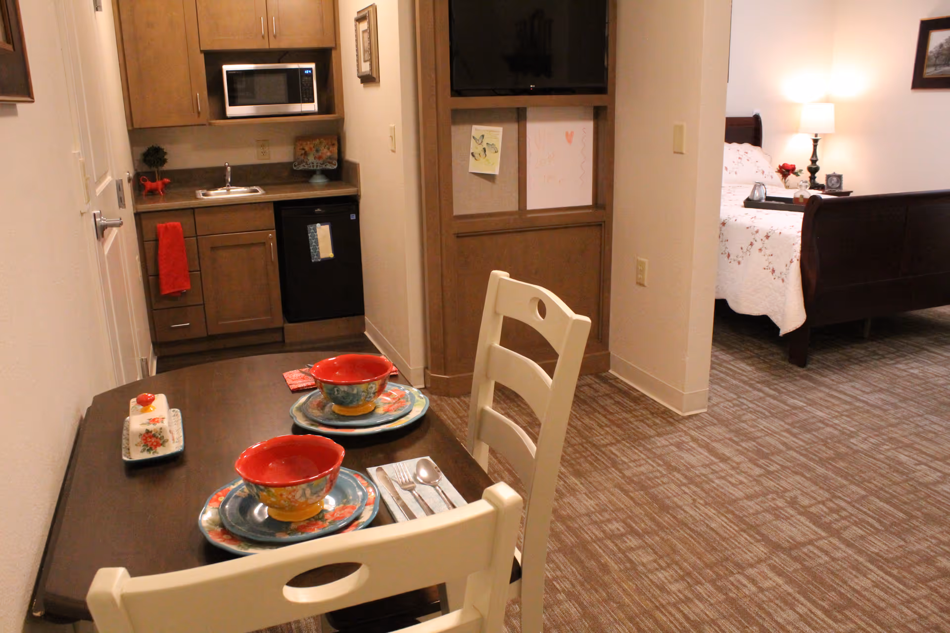 Apartment-style interior with a small kitchenette, a two-place dining table set with colorful dishes, and a partially visible bedroom.