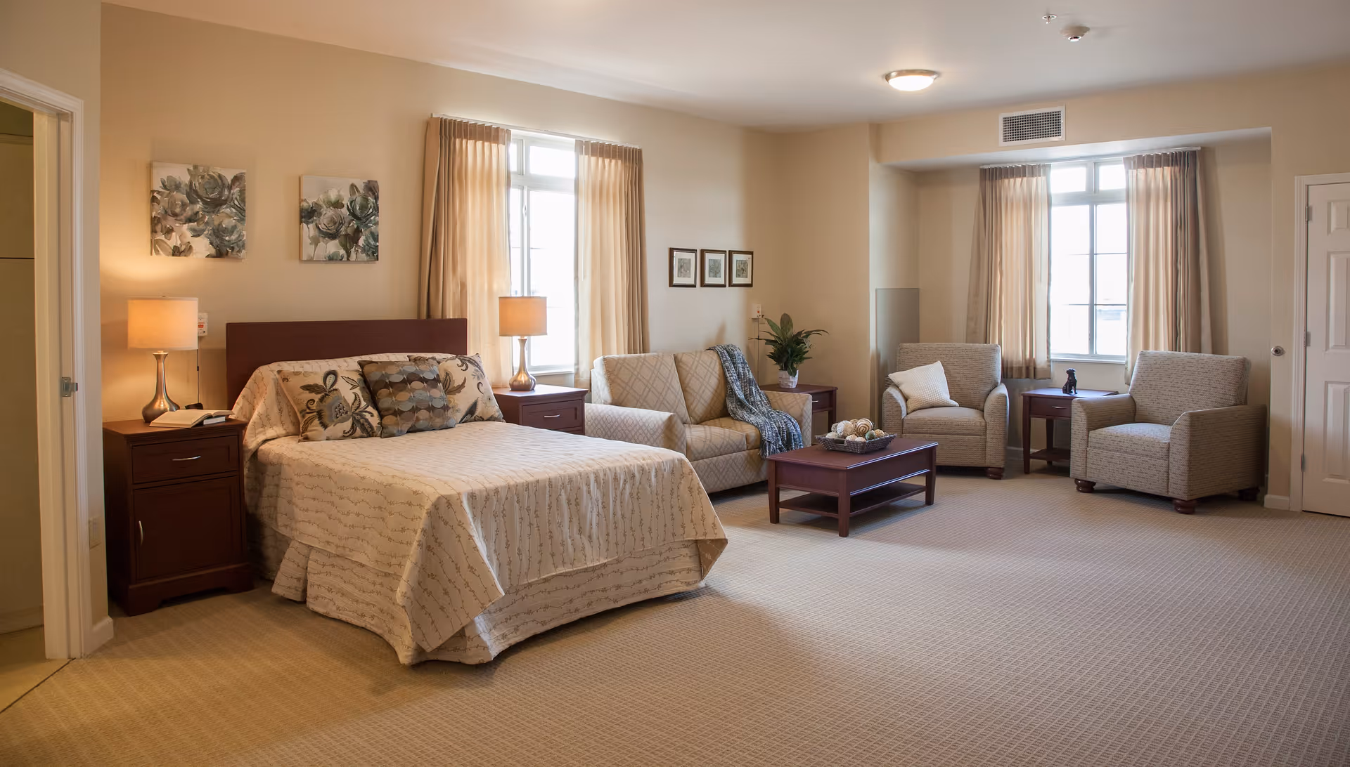 A spacious bedroom with a large bed covered in a beige patterned bedspread, two bedside tables with lamps, a loveseat, two armchairs, a coffee table, and windows with beige curtains letting in natural light. The walls are decorated with framed artwork and there is a door leading to another room.