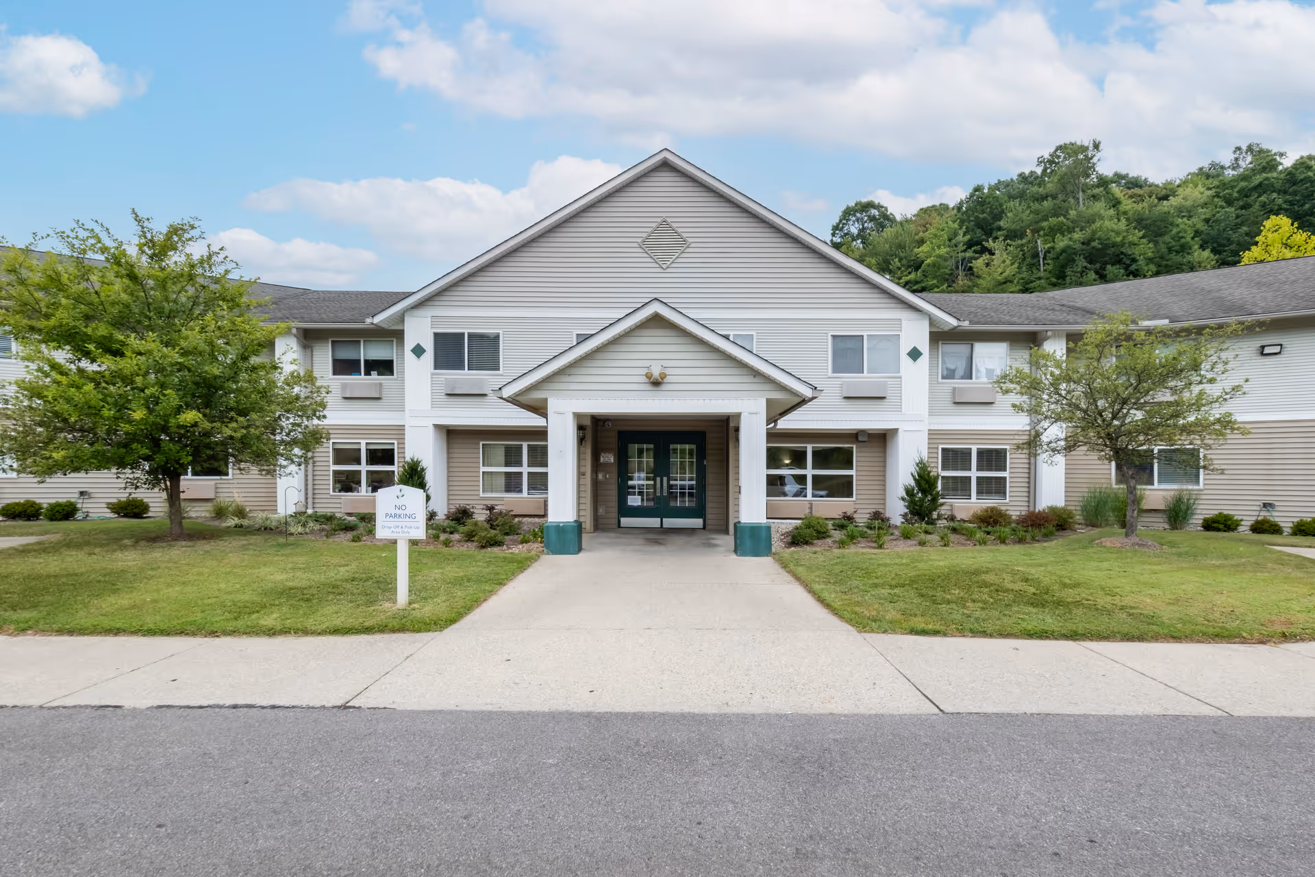 Front entrance of a two-story senior living facility with a covered porte-cochere, lawn, and trees.