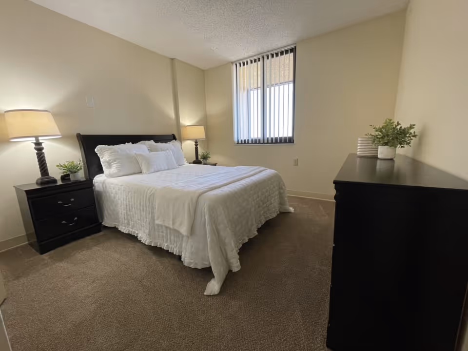 Well-lit bedroom with a bed dressed in white linens, two bedside tables with lamps, a dresser, and a window with vertical blinds.