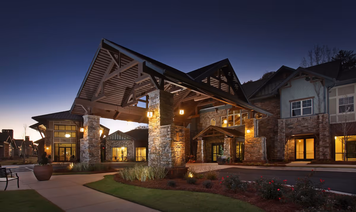 Dusk view of a senior living facility entrance with a large covered porte-cochère, stone pillars, and warm exterior lighting.