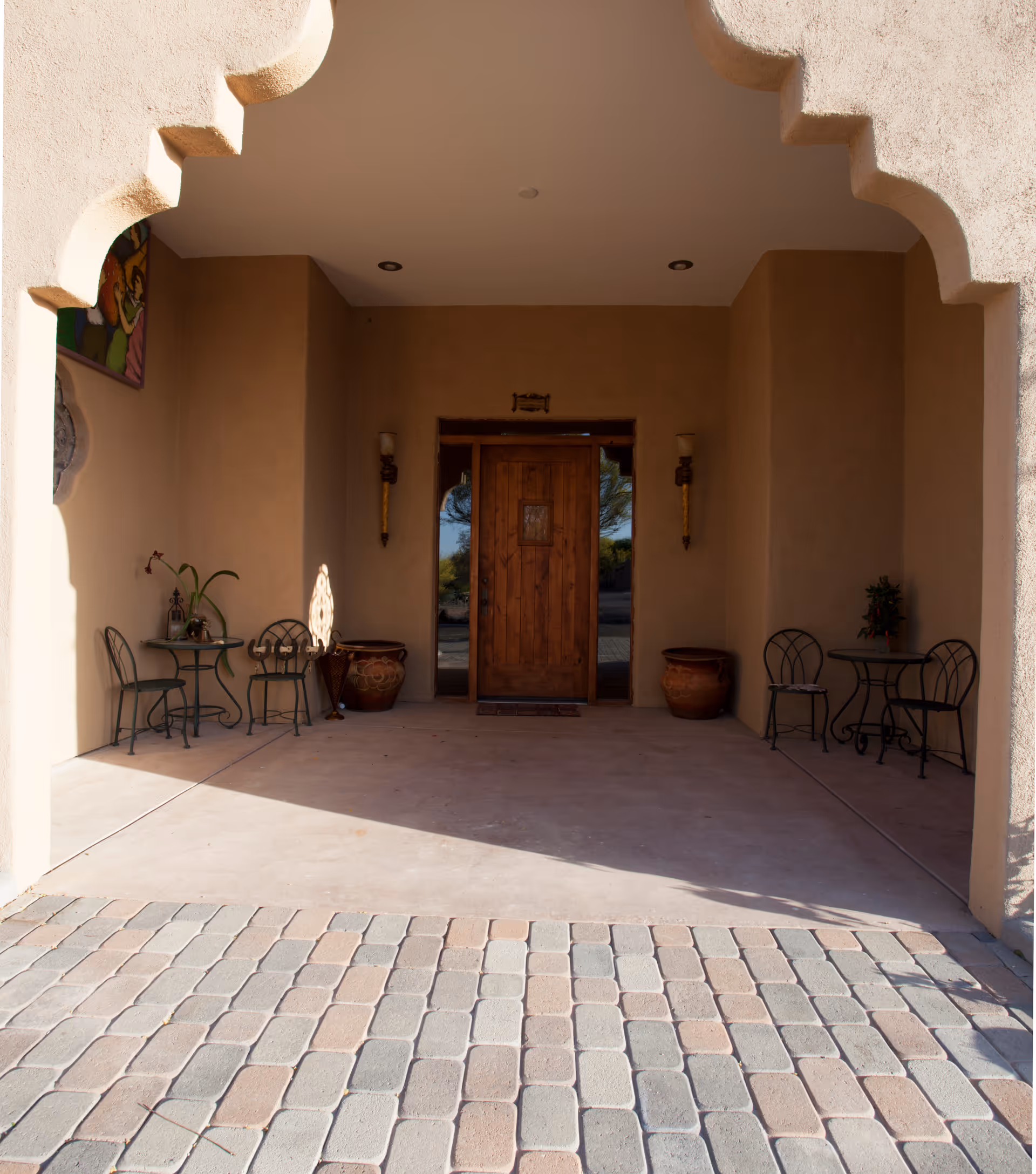 Recessed front entrance with a wooden door under a decorative archway flanked by small bistro tables, potted plants, and a paved driveway.