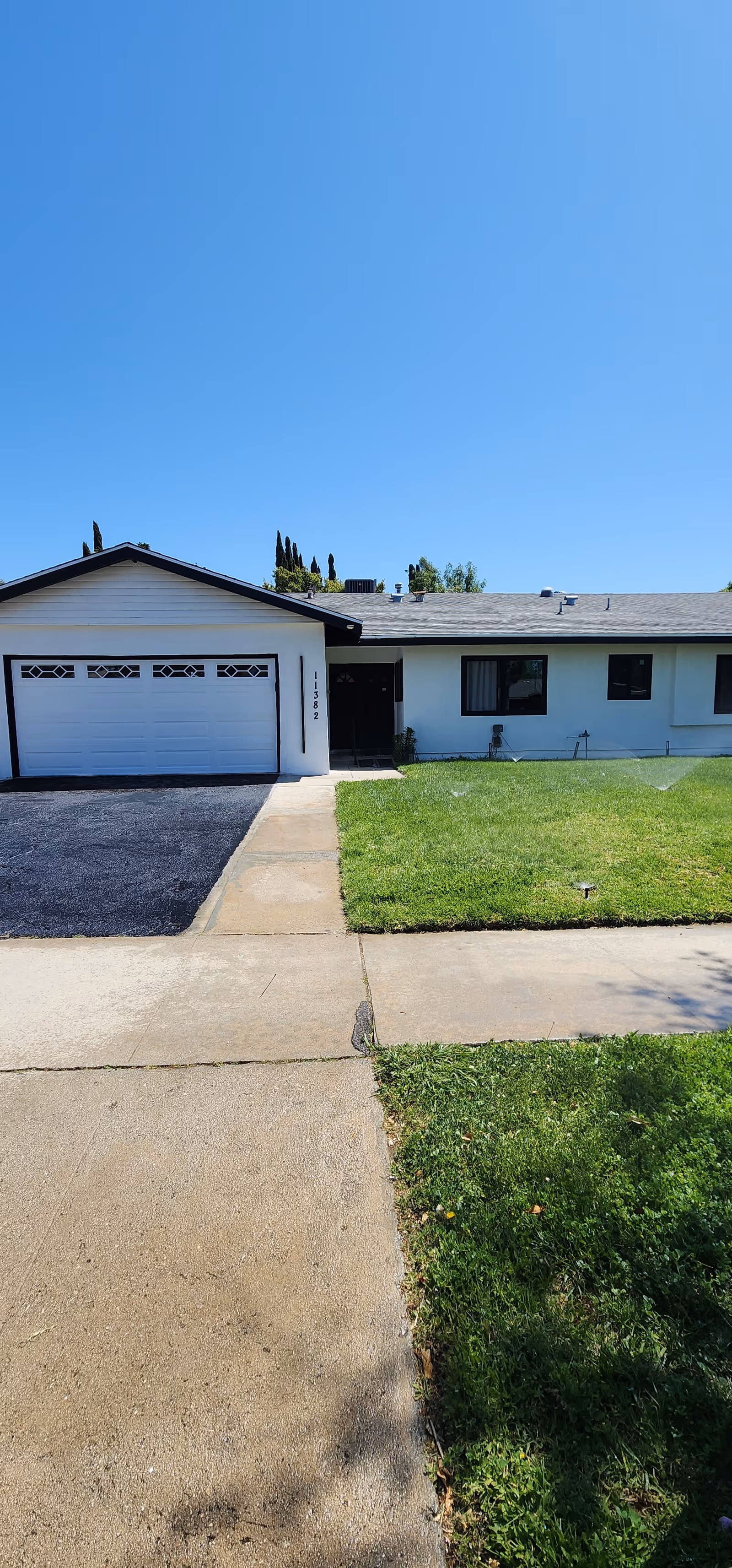 Front exterior view of a single-story house with a white garage door, a black driveway, a concrete walkway, and a green lawn under a clear blue sky.