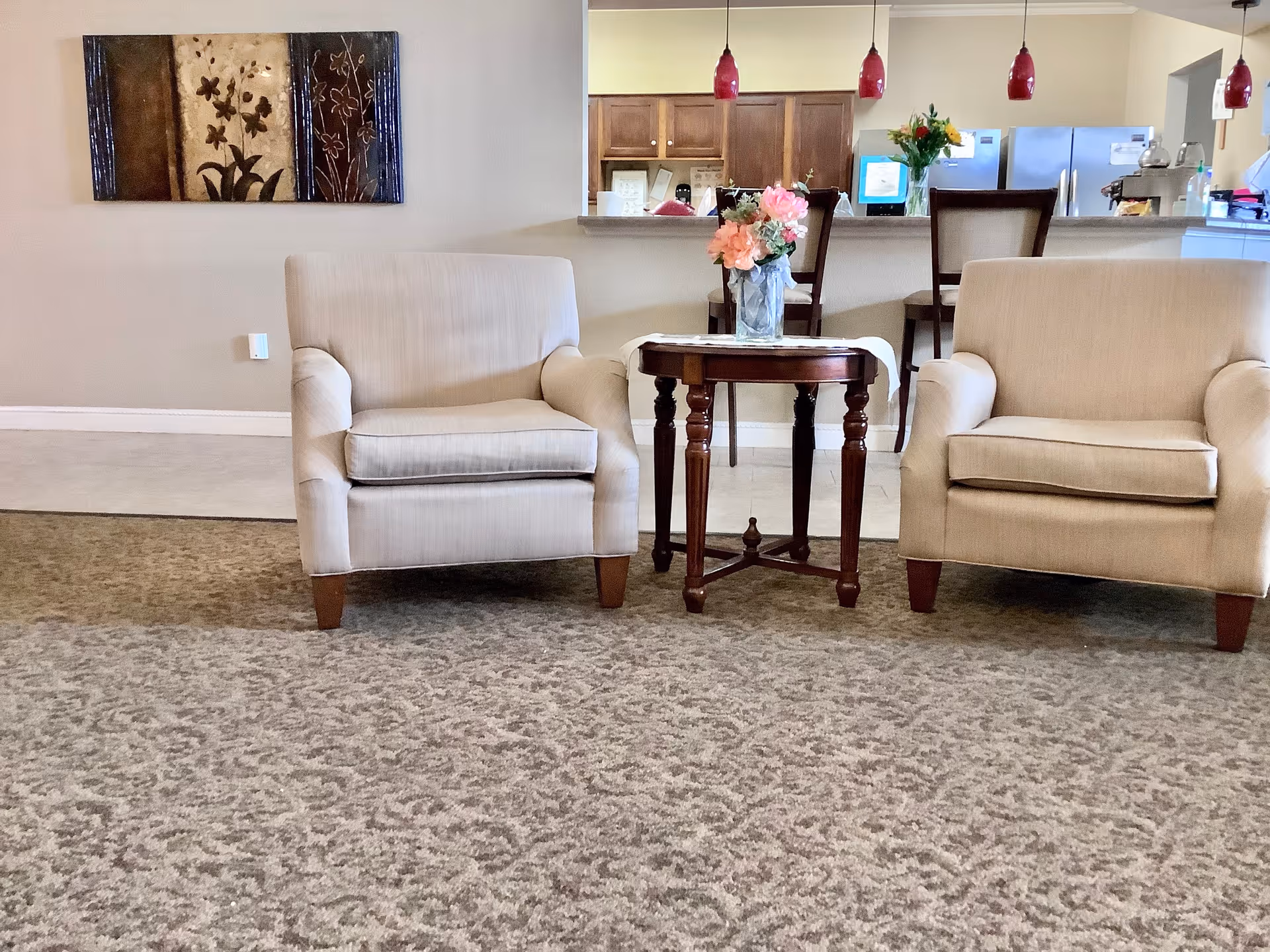 Two beige armchairs flank a small wooden side table with a vase of flowers in a carpeted common area with a view into a kitchen counter.