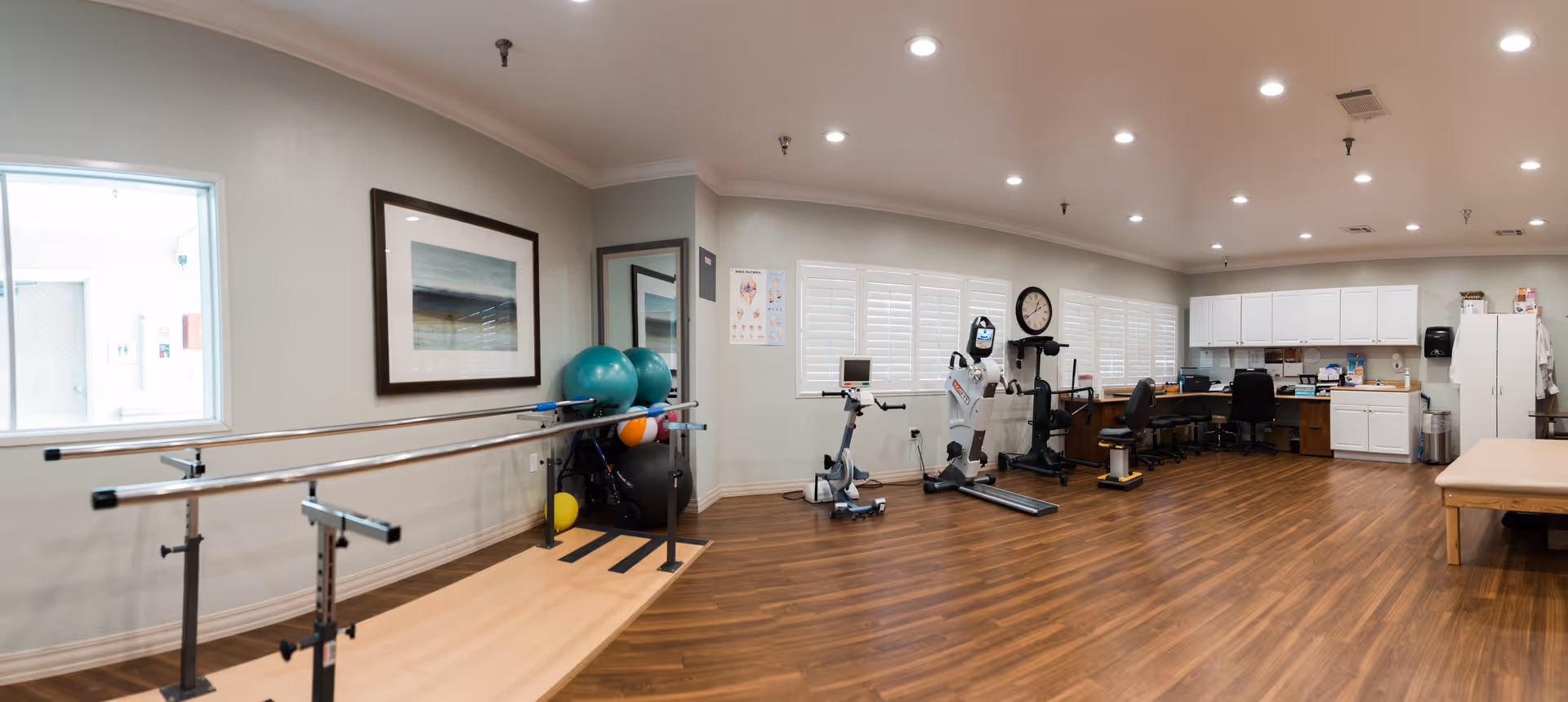 A spacious physical therapy or rehabilitation room with wooden flooring, parallel bars for walking exercises, exercise balls, stationary exercise equipment, a clock on the wall, and a desk area with chairs and cabinets. The room is well-lit with recessed ceiling lights and has large windows with white shutters.