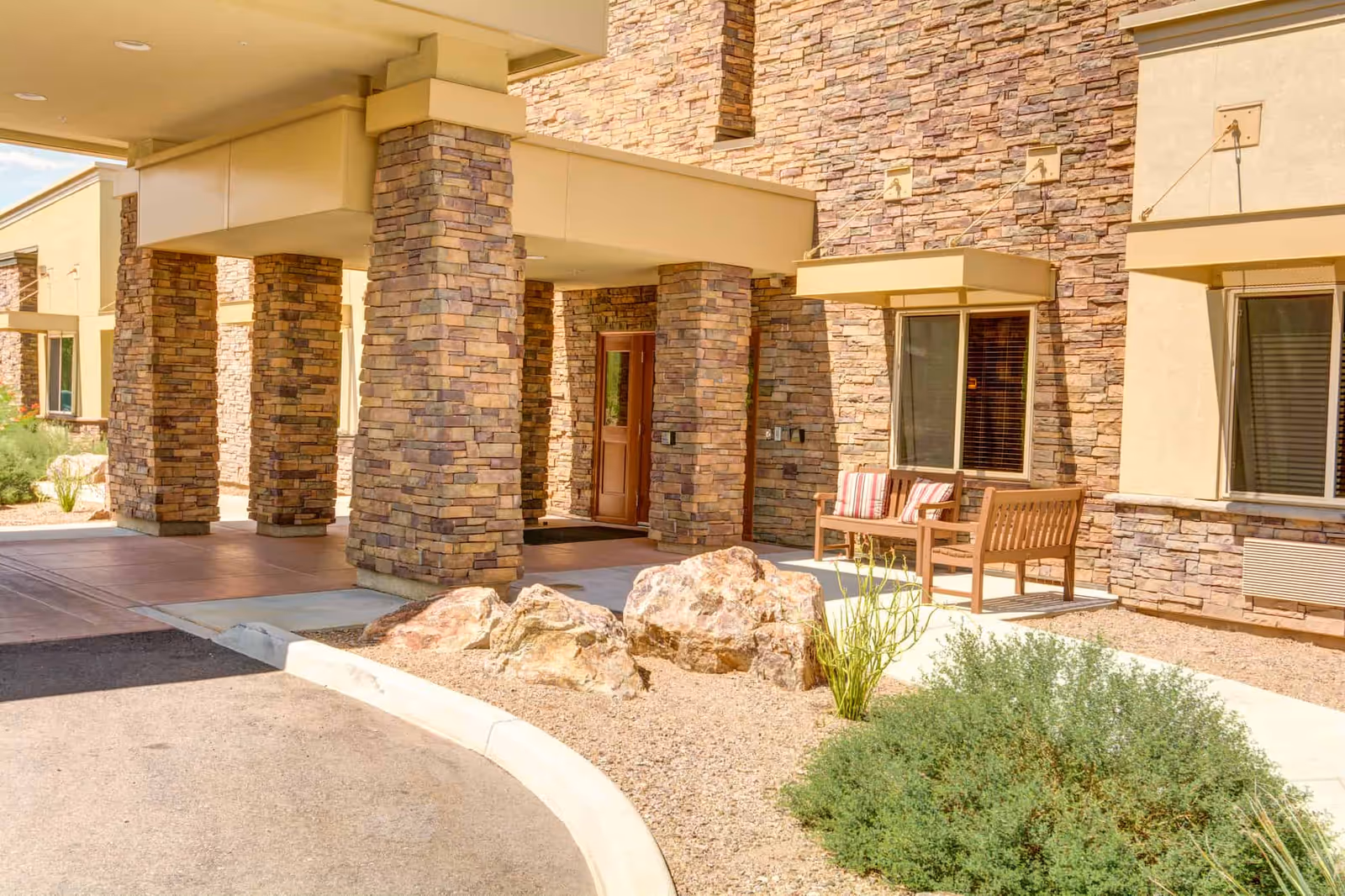 Entrance area of a building with stone pillars and walls, a covered driveway, a wooden bench with striped cushions, and desert landscaping with rocks and shrubs.