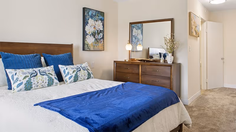 A neatly made bed with white bedding and a blue throw blanket in a senior living community bedroom. The bed has decorative pillows with peacock designs and solid blue pillows. A wooden dresser with a large mirror, two lamps, and decorative items is against the wall. There are two framed floral paintings on the walls, and a door is open leading to another room.
