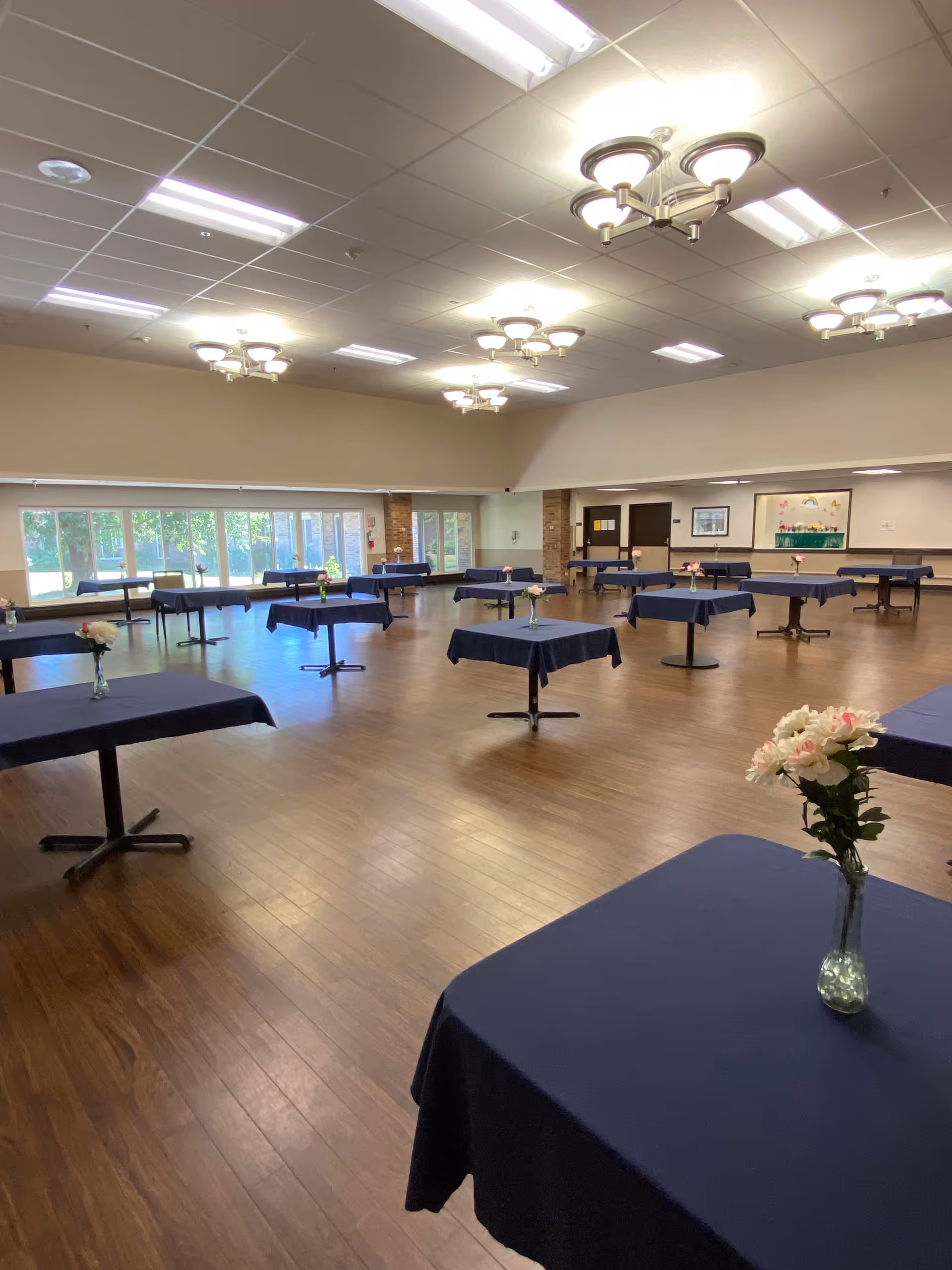 A spacious dining room with multiple square tables covered in dark blue tablecloths, each adorned with a small vase of flowers. The room has wooden flooring, large windows letting in natural light, and ceiling lights with multiple fixtures.