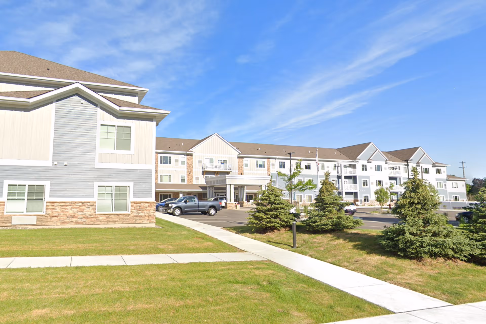 Front exterior of a three-story senior living building with a parking lot, small evergreen trees, a grassy lawn, and a sidewalk under a blue sky.