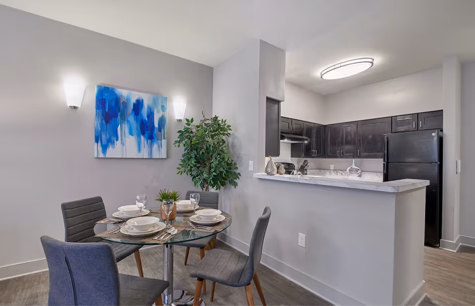 A modern dining area with a round glass table set for four, featuring white plates, bowls, and glasses. Four gray upholstered chairs surround the table. On the wall above the table is a blue abstract painting, flanked by two wall sconces. To the right is a kitchen with dark wood cabinets, a black refrigerator, and a marble countertop bar. A potted green plant is placed in the corner between the dining area and kitchen.