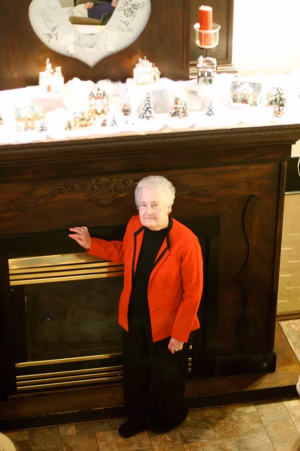 An elderly woman with white hair wearing a red jacket and black pants stands in front of a dark wooden fireplace mantel decorated with a miniature winter village scene and a red candle in a glass holder. A heart-shaped mirror is mounted above the mantel.