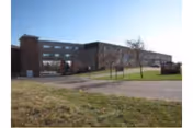 Brick multi-story retirement facility building with a lawn and driveway under a clear blue sky.